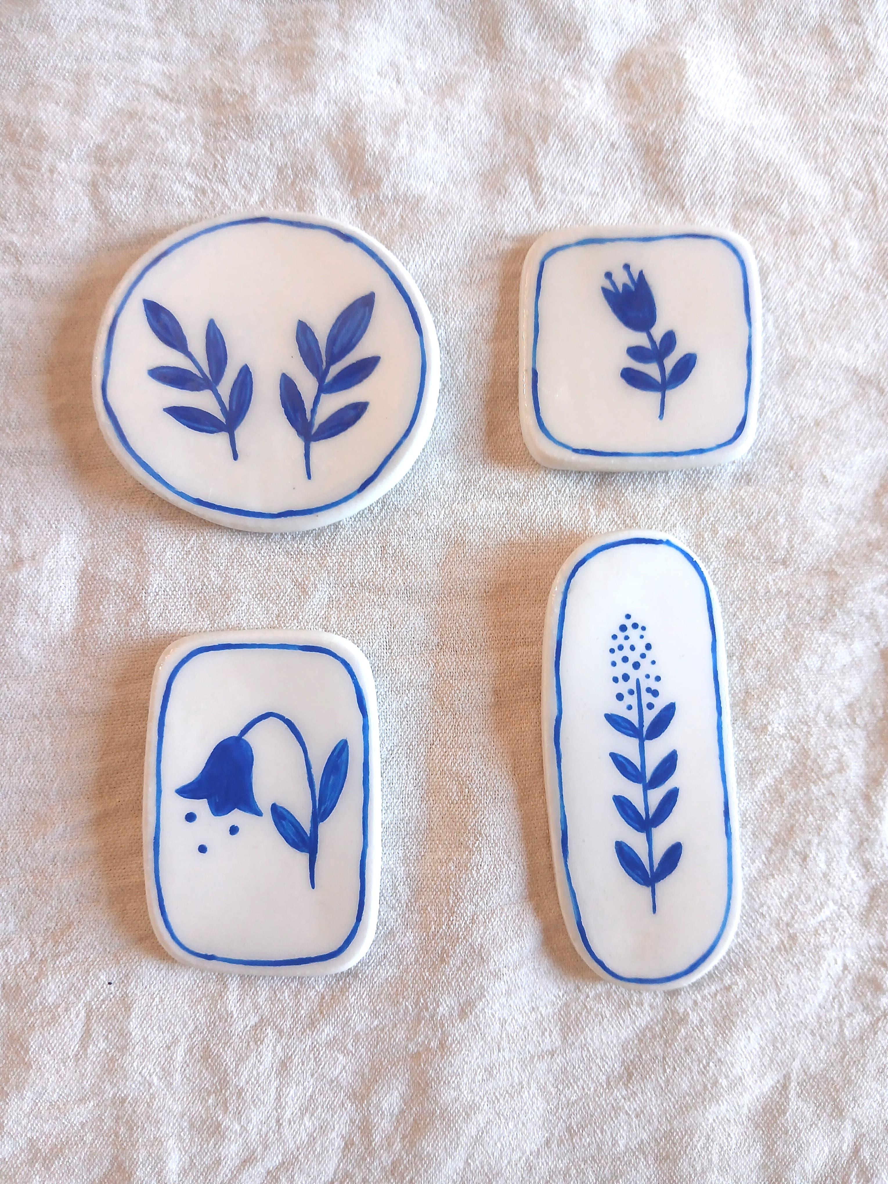 Quatre magnets botaniques en argile blanche peinte en bleu, posés sur lin